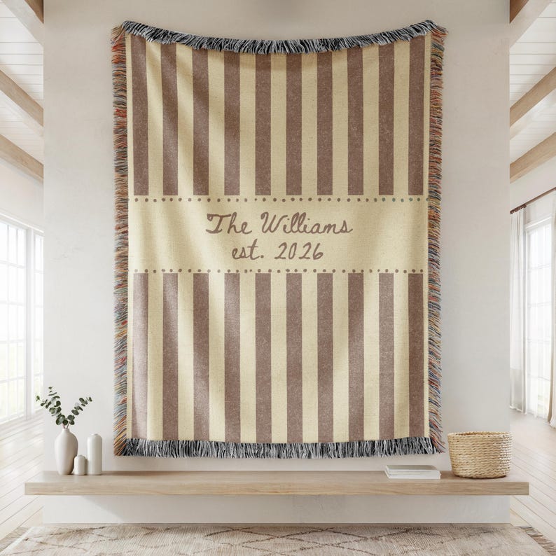 Puede incluir: Una manta a rayas beige y marr&oacute;n con un borde con flecos. La manta presenta el texto "The Williams est. 2026" en una fuente decorativa. La manta se exhibe en una estanter&iacute;a de madera.