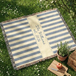 Puede incluir: Una manta de picnic rectangular con rayas azules y crema. El panel central presenta el texto "The Williams est. 2026". La manta tiene un borde con flecos y est&aacute; colocada sobre hierba. Tambi&eacute;n se ven una peque&ntilde;a planta en maceta, una tabla de cortar con una manzana y un cuchillo, y una cesta de picnic.