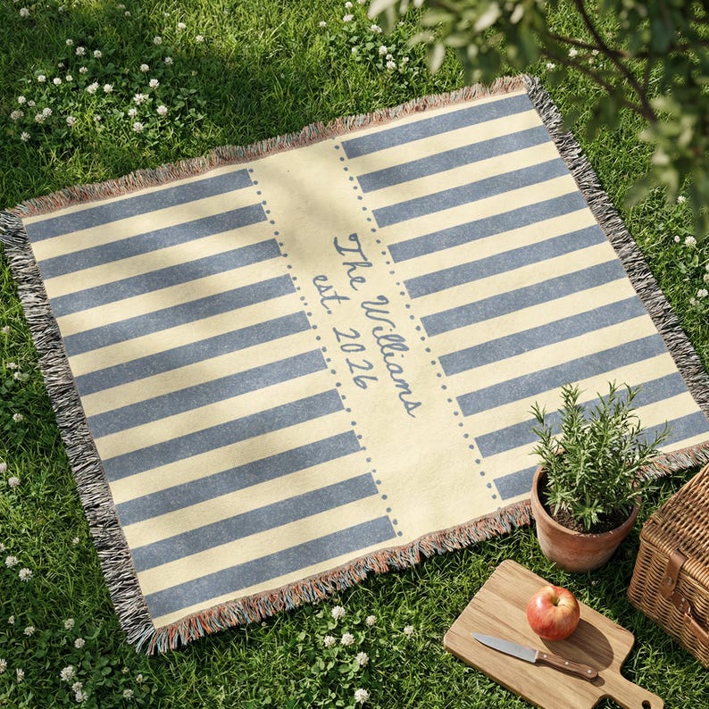 Puede incluir: Una manta de picnic rectangular con rayas azules y crema. El panel central presenta el texto "The Williams est. 2026". La manta tiene un borde con flecos y est&aacute; colocada sobre hierba. Tambi&eacute;n se ven una peque&ntilde;a planta en maceta, una tabla de cortar con una manzana y un cuchillo, y una cesta de picnic.