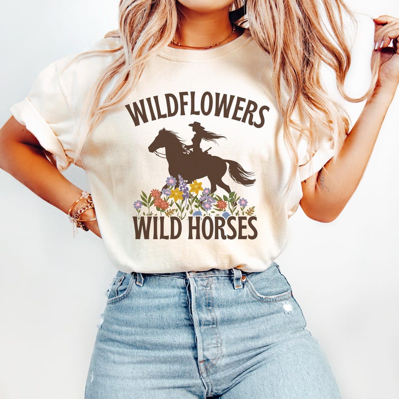May include: A white t-shirt with the text 'Wildflowers Wild Horses' in brown text. A silhouette of a woman riding a horse is between the words. The horse and woman are brown and the background is white. The shirt is being worn by a person with long blonde hair.