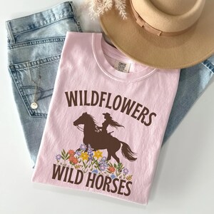 May include: Pink t-shirt with a brown silhouette of a woman riding a horse with the text "Wildflowers Wild Horses" printed on it.