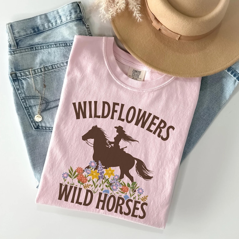 May include: Pink t-shirt with a brown silhouette of a woman riding a horse with the text "Wildflowers Wild Horses" printed on it.