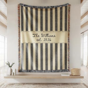 Puede incluir: Una manta personalizada con rayas verticales grises sobre un fondo crema. El centro presenta el texto "The Williams est. 2026" en una fuente de escritura. La manta tiene bordes con flecos y se muestra en una pared.