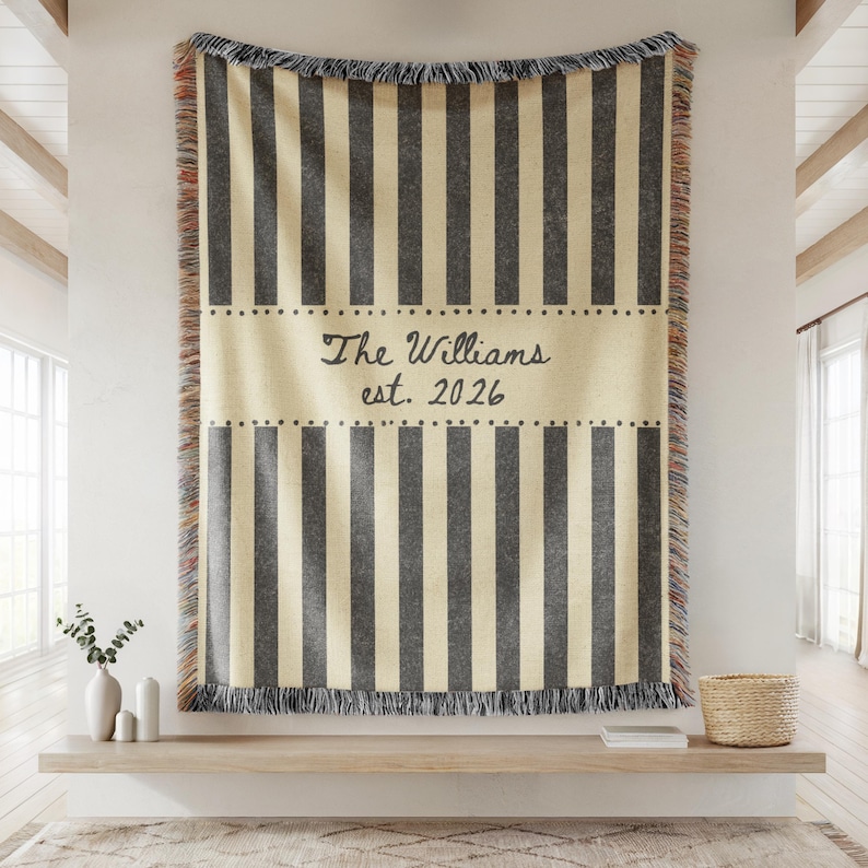 Puede incluir: Una manta personalizada con rayas verticales grises sobre un fondo crema. El centro presenta el texto "The Williams est. 2026" en una fuente de escritura. La manta tiene bordes con flecos y se muestra en una pared.
