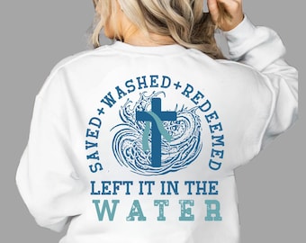 May include: White sweatshirt with a blue cross design, surrounded by the words "SAVED + WASHED + REDEEMED". Below the design, the text reads "LEFT IT IN THE WATER". The design features a cross with a blue ribbon.