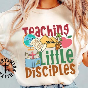 Könnte beinhalten: Cremefarbenes T-Shirt mit den Worten "Teaching Little Disciples" in Rot, Grün und Braun. Das Design umfasst einen Globus, Pyramiden, eine Schriftrolle, ein Kreuz, ein Buch und einen herzförmigen Apfel. Das Shirt hat eine lässige, entspannte Passform.
