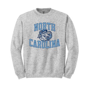 May include: A gray sweatshirt with a blue and white North Carolina Tar Heels logo. The logo features a mascot with a sailor hat and the text "UNC" on it. The text "NORTH CAROLINA" is printed in a blue arch above the mascot.