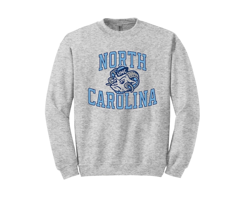 May include: A gray sweatshirt with a blue and white North Carolina Tar Heels logo. The logo features a mascot with a sailor hat and the text "UNC" on it. The text "NORTH CAROLINA" is printed in a blue arch above the mascot.