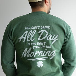 May include: Green sweatshirt with white text that reads "YOU CAN'T DRINK ALL DAY IF YOU DON'T START IN THE MORNING" and a four-leaf clover graphic. A beige and green baseball cap is worn.