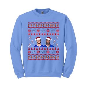May include: A light blue sweatshirt with a festive design. The design includes images of two people wearing Santa hats, candy canes, snowflakes, and other holiday-themed elements. The sweatshirt is a long-sleeved crew neck.