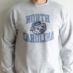 May include: A gray sweatshirt with a blue and white North Carolina logo. The logo features a ram wearing a UNC hat.