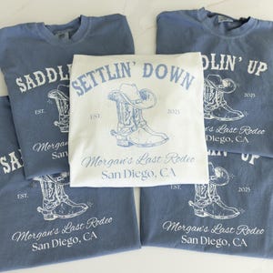 May include: A collection of blue and white t-shirts with cowboy-themed graphics. The shirts feature a cowboy boot and hat design, along with the text "Saddlin' Up", "Settlin' Down", and "Morgan's Last Rodeo San Diego, CA".