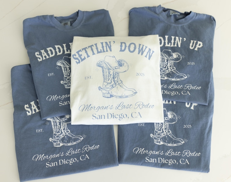 May include: A collection of blue and white t-shirts with cowboy-themed graphics. The shirts feature a cowboy boot and hat design, along with the text "Saddlin' Up", "Settlin' Down", and "Morgan's Last Rodeo San Diego, CA".