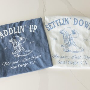 May include: Two t-shirts, one blue and one white, with cowboy boot and hat graphics. The blue shirt says "SADDLIN' UP" and the white shirt says "SETTLIN' DOWN". Both shirts have "Morgan's Last Rodeo, San Diego, CA" printed below.