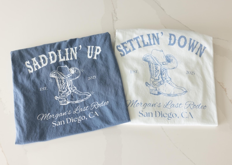 May include: Two t-shirts, one blue and one white, with cowboy boot and hat graphics. The blue shirt says "SADDLIN' UP" and the white shirt says "SETTLIN' DOWN". Both shirts have "Morgan's Last Rodeo, San Diego, CA" printed below.