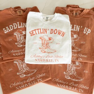 May include: A collection of t-shirts in shades of brown and white, featuring rodeo-themed designs. The shirts display text such as "Saddlin' Up" and "Settlin' Down", along with cowboy boot and hat illustrations. The text "Ashley's Last Rodeo, Nashville, TN" is also visible.