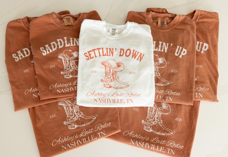 May include: A collection of t-shirts in shades of brown and white, featuring rodeo-themed designs. The shirts display text such as "Saddlin' Up" and "Settlin' Down", along with cowboy boot and hat illustrations. The text "Ashley's Last Rodeo, Nashville, TN" is also visible.