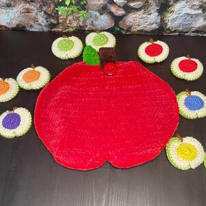 May include: A handmade, red apple-shaped trivet with a green leaf and brown stem, surrounded by crocheted coasters in various colors. The coasters feature orange, purple, yellow, red, green, and blue centers with white and green borders.