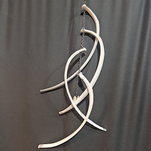 May include: A hanging mobile sculpture composed of four curved, white, bone-like segments suspended vertically from a thin, dark metal chain. The segments are arranged in an overlapping, asymmetrical pattern, creating a layered structure.