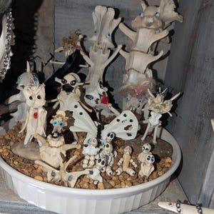 May include: A white ceramic bowl filled with small white bone sculptures of various creatures, including butterflies, skeletons, and humanoid figures. The sculptures have blue eyes and are arranged on a bed of small brown stones.