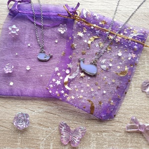 May include: Two silver necklaces with purple whale charms on a purple background. The necklaces are displayed on a purple and gold fabric with a star and moon pattern.