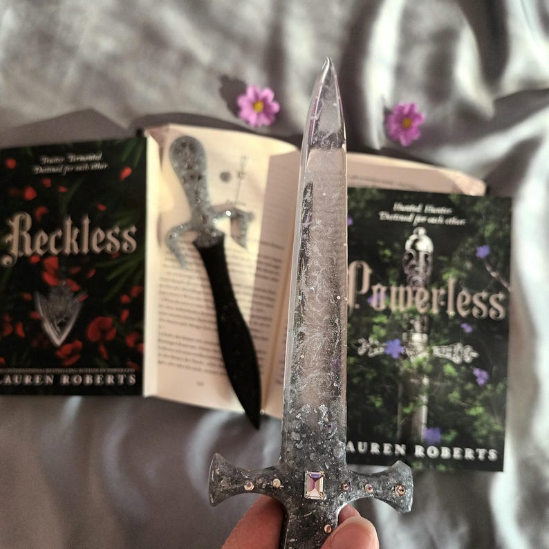 Puede incluir: Dos novelas de fantas&iacute;a, "Reckless" y "Powerless" de Lauren Roberts, con un marcap&aacute;ginas de espada de brillo plateado. El marcap&aacute;ginas de espada tiene un mango transparente y una hoja plateada con brillo incrustado en la resina.