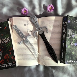 Puede incluir: Dos libros de bolsillo de Lauren Roberts, "Reckless" y "Powerless", con el texto "Truiter. Tirmented Destined for each other" y "Hunted Hunter Destined for each other" en las portadas. Los libros est&aacute;n abiertos y tienen dos marcadores de daga decorativos plateados y negros.