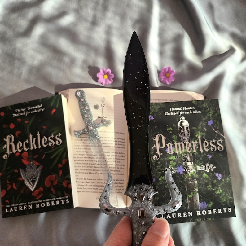 Puede incluir: Dos libros de bolsillo titulados "Reckless" y "Powerless" de Lauren Roberts. Los libros est&aacute;n abiertos y una daga plateada y negra con pedrer&iacute;a est&aacute; colocada entre las p&aacute;ginas. Los libros tienen un fondo oscuro con flores rojas y moradas.