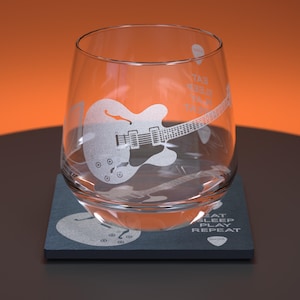 May include: A clear glass tumbler with an etched design of a guitar and the text "Eat Sleep Play Repeat". The tumbler is sitting on a dark blue coaster with the same design.