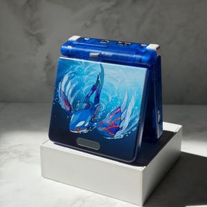 Puede incluir: Consola de juegos portátil azul con un diseño de Kyogre en la parte delantera. La consola está colocada sobre una caja blanca. El diseño de Kyogre presenta tonos de azul y rojo sobre un fondo azul oscuro con efectos de agua.