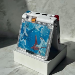 May include: A white and clear handheld gaming device with a screen showing two cartoon characters against a blue sky. The device has red and blue accents and is on a white box.
