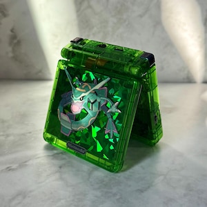 May include: A transparent green handheld gaming console with a holographic design featuring a mythical creature. The console is open, revealing the screen and the Nintendo logo. The design has a green and pink color scheme.