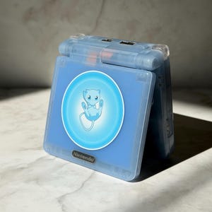 May include: A light blue Nintendo Game Boy Advance SP console. The console is open, showing a screen with a circular graphic of the Pokémon Mew. The Nintendo logo is visible below the screen.