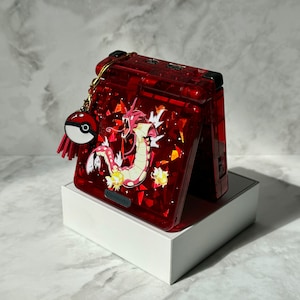 Puede incluir: Consola de juegos portátil roja con un diseño de criatura dragón y el logotipo de Nintendo. La consola está decorada con purpurina roja y un llavero con un charm de Pokéball. La consola está sobre una caja blanca.