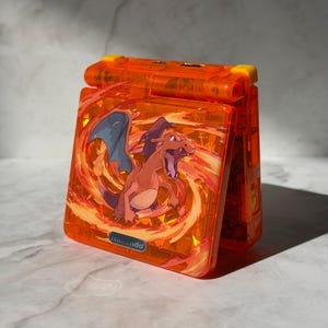 Może przedstawiać: Pomarańczowe etui na Game Boy Advance SP z grafiką Charizarda. Obudowa ma przezroczysty design z żółtym zawiasem i logo Nintendo. Ilustracja Charizarda jest umieszczona na ognistym tle.