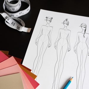 May include: A white sheet of paper with three fashion design templates of women. The paper is on a black surface with a blue pencil, a measuring tape, and a stack of colorful fabric swatches.