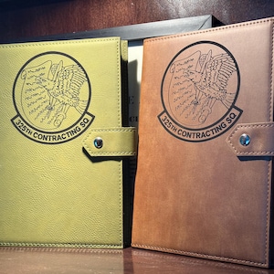 May include: Two rectangular notebooks with snap closures, one in olive green and one in brown. Each cover features a black circular emblem with an eagle and the text 325TH CONTRACTING SQ. The covers have a textured finish and visible stitching along the edges.