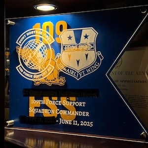 Może przedstawiać: Niebiesko-złota tablica z emblematem 109th Force Support Squadron. Tablica zawiera tekst, taki jak "SUNT NOSTRAE VIRES" i "109th Airlift Wing". Wyświetla również imię "Lt Col Ashley N Fitzgibbon" oraz daty "Sept 11, 2021 - June 11, 2025."