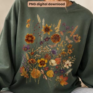 May include: A dark olive green crewneck sweatshirt featuring a central graphic of various wildflowers, wheat stalks, and autumn leaves in shades of orange, yellow, and purple. The text "PNG digital download" is printed in a small box at the top.