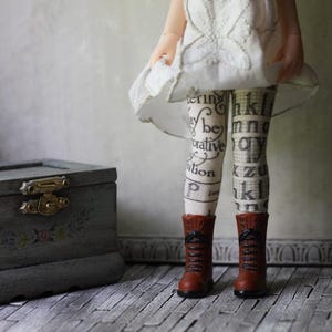 May include: A doll in a white dress and patterned tights with text, wearing brown boots. A grey wooden box with a gold clasp and floral details is on the left. The background is a neutral grey.