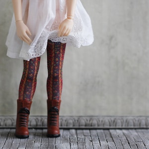 Könnte beinhalten: Eine Puppe trägt ein weißes Kleid mit Spitzenbesatz und gemusterten Strumpfhosen. Die Puppe trägt braune Stiefel und steht auf einem Holzboden.