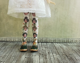 May include: A pair of white tights with a pattern of women's faces and floral accents. The tights are worn by a doll with a white dress and are paired with dark green shoes.