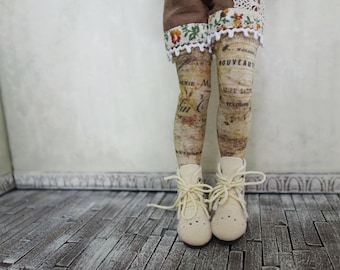 May include: A doll's legs are shown wearing beige boots with cream-colored laces. The doll is also wearing patterned stockings with the word "NOUVEAU" printed on them. The doll's shorts are brown with floral and lace trim.