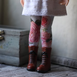 Pode incluir: Uma boneca vestindo um vestido branco com renda e meias com padrão floral. A boneca está usando botas marrons com cadarços.
