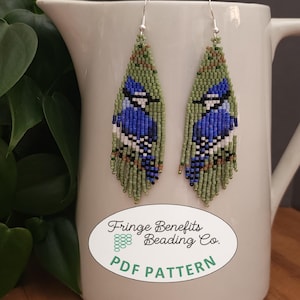 May include: A pair of beaded earrings featuring a blue jay design with green, blue, and white beads. The earrings are hanging from silver hooks. The image also includes a white pitcher and a green plant.
