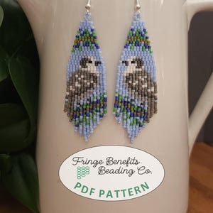May include: Handmade beaded earrings with a blue, green, and gray color scheme. The earrings feature a design of a small animal. The earrings are hanging on a white pitcher. The text on the pitcher says "Fringe Benefits Beading Co. PDF PATTERN."