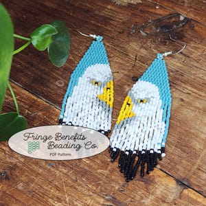 Könnte beinhalten: Ein Paar Perlenohrringe mit einem weißen Adler-Design mit blauen, gelben und grauen Akzenten. Die Ohrringe haben schwarze Fransen und silberne Haken. Die Ohrringe sind mit "Fringe Benefits Beading Co. PDF Pattern" gekennzeichnet.