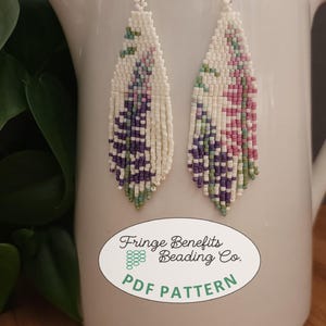 May include: A pair of beaded earrings with a floral design. The earrings are white with purple, pink, and green accents. The earrings have a fringe at the bottom and are hanging from silver hooks. The earrings are displayed on a white jug.