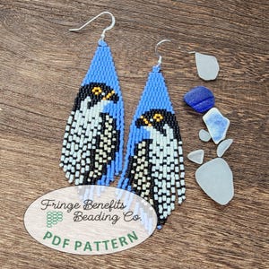 May include: A pair of beaded earrings with a bird design. The earrings feature a blue background with a black, white, and yellow bird design. The earrings have a fringe at the bottom and are hanging on a silver hook. The earrings are on a wooden surface.