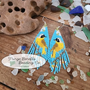 May include: A pair of blue and yellow beaded earrings with a bird design. The earrings are hanging from silver hooks and have a fringe of beads. The earrings are on a wooden surface with small pieces of glass.